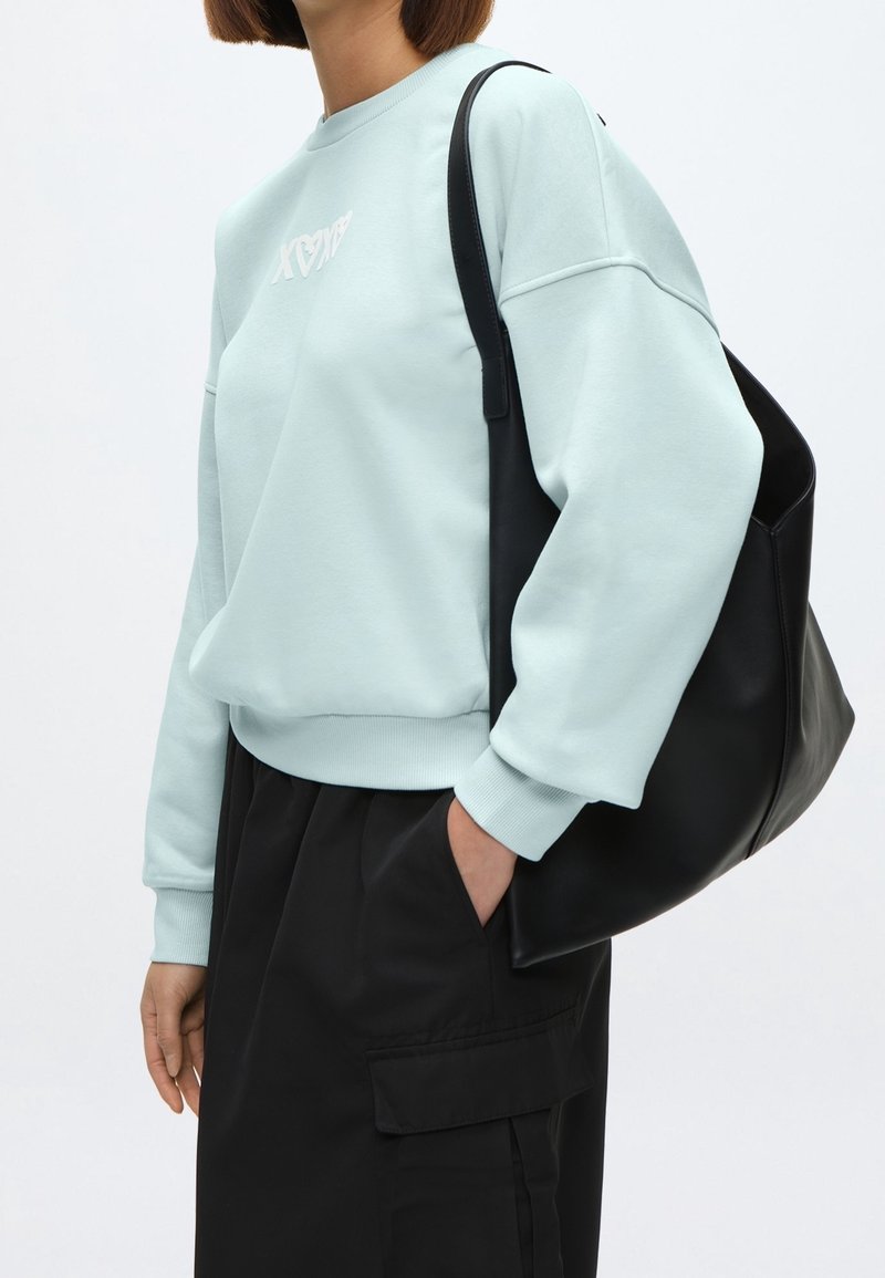 Personne portant un sweat-shirt bleu clair et un pantalon noir, la main dans la poche, portant un grand sac noir à l'épaule sur fond blanc.