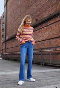 Stripet strikkegenser i oransje, rosa og blå med høyt hals; parret med blå utsvingte jeans og hvite joggesko; bakgrunn av tredekket.