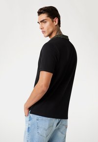 Black polo shirt with a textured green collar, short sleeves, and a relaxed fit, paired with light blue jeans.