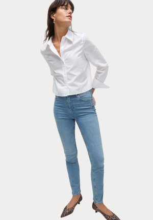 Camisa blanca de botones, ajustada, con cuello y mangas largas; combinada con pantalones vaqueros ajustados de color azul claro y bailarinas de punta con estampado de leopardo.