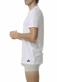 Biała bawełniana koszulka T-shirt z krótkim rękawem, z czarnym logo Adidas na dole po lewej stronie. W połączeniu z szarymi szortami. Widok z boku.