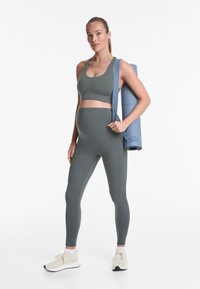 Sötétzöld sportmelltartó és leggings, sima, rugalmas anyagból készült. Bézs sportcipőt visel, és egy kék jóga matra tart.