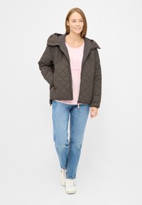Gequilted donkerbruine jas met een afneembare capuchon, gedragen over een lichtroze t-shirt, gecombineerd met blauwe jeans en witte sneakers.