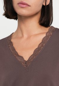 Top marrón de cuello en V con detalles intrincados de encaje a lo largo del escote, textura suave y diseño ajustado.