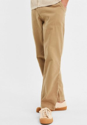 Beige cotton pants with a relaxed fit, featuring side pockets and subtle stitching details. Worn with light-colored sneakers.