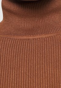 Ribbetu turtleneck-neule lämpimässä ruskeassa. Pehmeä tekstuuri hienoine pystysuorine viivoineen ja tiukka istuvuus kaulassa. Ei näkyvää varustusta tai koristeita.
