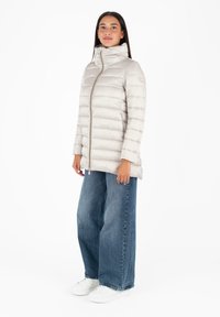 Lichtbeige pufferjack met hoge kraag, ritssluiting en horizontale stiksels. Gecombineerd met wijde blauwe jeans en witte sneakers.