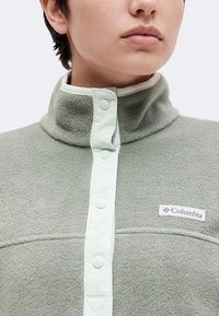 Άτομο που φοράει ένα ανοιχτοπράσινο fleece pullover Columbia με ψηλό γιακά και κλείσιμο με κουμπιά τύπου σναπ.