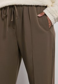 Närbild av bruna dragsko-byxor med en hand i fickan och en ärm av en krämfärgad stickad tröja synlig.