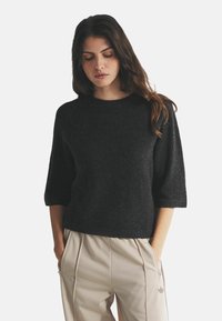 Juoda tekstūruota megztinis su apvalia iškirpte ir trijų ketvirčių rankovėmis, derinamas su šviesiai rudais, raukšlėtais kelnėmis su šoninėmis kišenėmis.