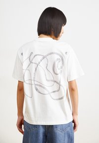 Hvid bomulds t-shirt med et sølvreb design på ryggen, afslappet pasform, korte ærmer og rund halsudskæring. Denim shorts synlige.