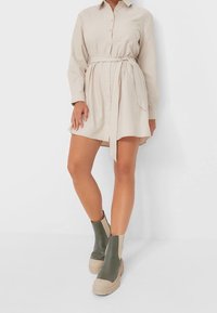 Beige skjortklänning med knytmidja, långa ärmar och knappstängning, i kombination med gröna och beige ankelskor med en texturerad sula.