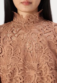 Haut en dentelle avec des motifs floraux complexes de couleur pêche douce. Comprend un col haut et une broderie détaillée sur un tissu transparent.