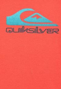 Korálové tričko s modrou vlnou a černým logem "Quiksilver". Měkká, hladká textura s uvolněným, moderním designem.