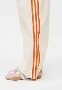 Εκρού παντελόνια με τρεις κάθετες πορτοκαλί ρίγες, φορεμένα πάνω από ροζ και λευκά παπούτσια μπαλέτου, σε ανοιχτό φόντο.