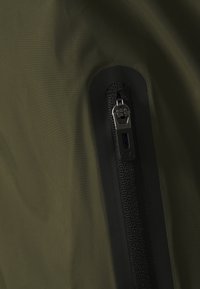 Olivgrön tyg med en jämn yta, med en svart dragkedja med en texturerad dragknapp och en förstärkt dragkedjeomkrets.