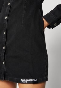 Černé denimové šaty s knoflíkovým zapínáním, dvěma bočními kapsami a dlouhými rukávy. Na spodním lemu je umístěna logo nášivka s nápisem "KARL LAGERFELD JEANS".
