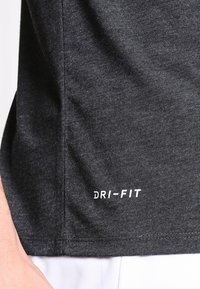 Ciemnoszara tkanina sportowa z widocznymi szwami i białym logo "DRI-FIT" blisko brzegu, częściowo pokazująca nadgarstek osoby oraz białe szorty.