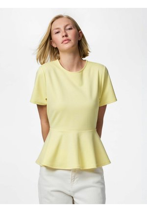 Jeune femme aux cheveux blonds mi-longs, vêtue d'un haut peplum jaune pâle à manches courtes et d'un pantalon blanc, debout sur un fond blanc.