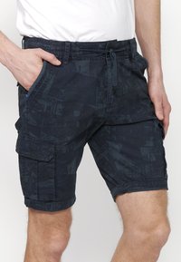 Sötét mintás cargo sort viselő férfi zsebekkel, húzózsinórral és felgombolt szegélyekkel, állva, keze a zsebében.