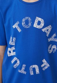 T-shirt en coton bleu avec un graphique circulaire blanc comportant le texte "L'AVENIR D'AUJOURD'HUI" dans différentes orientations et un effet vieilli.