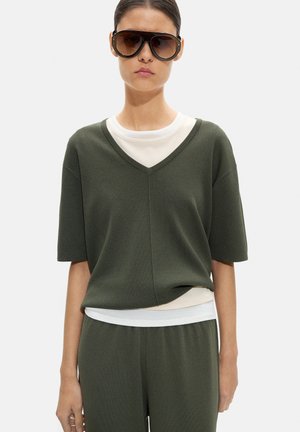 Kobieta w dużych ciemnych okularach przeciwsłonecznych, oliwkowozielonej bluzce z krótkim rękawem i dekoltem w serek, na białej koszuli oraz dopasowanych oliwkowozielonych spodniach, stojąca prosto.