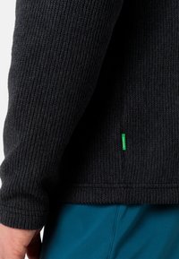 Pleteni pulover v temno sivi barvi z rebrasto teksturo. Ima zeleno oznako na robu in oprijete manšete. Usklajen z modrimi hlačami.