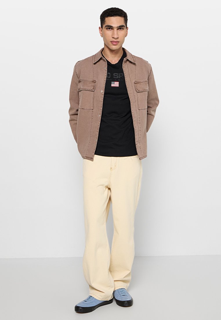 Giacca camicia in cotone marrone sopra una t-shirt grafica nera, abbinata a pantaloni giallo chiaro e scarpe blu. Quattro tasche sulla giacca.