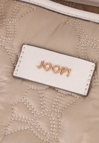 Beige kviltat tyg med relieferat blommönster. Vit etikett med guldtexten "JOOP!" sydd på, med texturerad yta och sömnad.