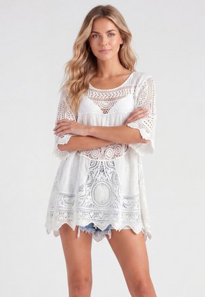 Kvinde står med armene over kors, iført en hvid blondetunika og denimshorts mod en lys, ensfarvet baggrund.