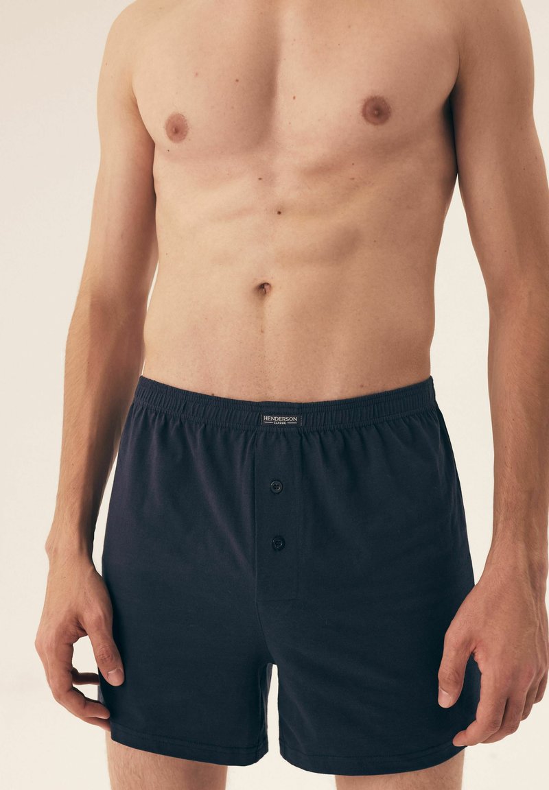 Czarne bawełniane bokserki z guzikiem, elastycznym pasem i gładką fakturą, zaprezentowane na modelu bez koszulki.