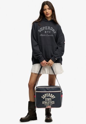 Ung kvinde iført en sort Superdry hættetrøje og hvid nederdel, der holder en navy Superdry State Athletics taske og står i sorte støvler.