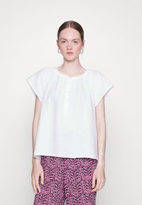 Top branco de manga curta com decote redondo e abertura com botões. Tecido leve com um acabamento texturizado. Combinado com calças cor-de-rosa estampadas.