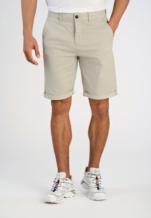 Mand iført beige mønstrede shorts med opsmøgede kanter og hvide træningssko, stående mod en hvid baggrund.