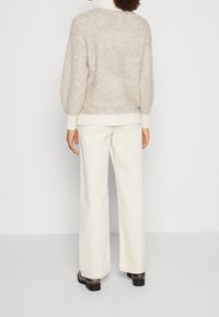 Beige, strukturierter Strickpullover mit Stehkragen und gerippten Bündchen, kombiniert mit weiten, cremefarbenen Hosen und schwarzen Schuhen. Rückansicht gezeigt.