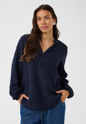 Femme aux longs cheveux bruns portant un chemisier bleu marine à manches longues et un jean bleu, debout les mains dans les poches, souriant doucement.