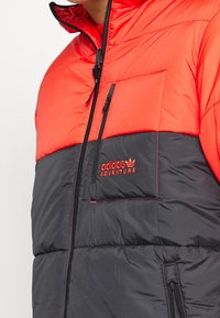 Puffjacka med röd övre del och svart undre del. Har en dragkedjestängning och en bröstficka med texten "adidas ADVENTURE" i röd.