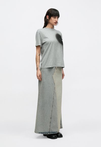 Tätoveeringutega naine, kes kannab lahtist halli t-särki, kahetoonilist pikka teksaseelikut ja musti platvormkingi, seisab valge ühevärvilise tausta ees.