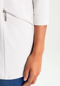 Veste en tissu gris clair et lisse avec des poignets repliés et une poche zippée sur le côté. Gros plan sur une main tenant la veste.