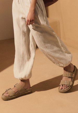 Person wearing beige suede sandals with buckles and loose, light-colored pants, standing on a tan floor with shadow.