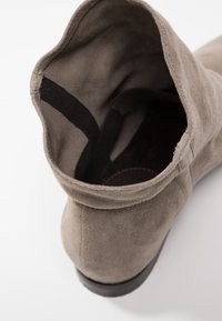 Bottine en daim taupe avec languette noire, coutures visibles et ouverture supérieure arrondie sur fond blanc.