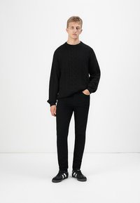 Giovane uomo con capelli biondi corti, che indossa un maglione nero a trecce, pantaloni neri e sneaker nere con strisce bianche, in piedi con una mano in tasca.