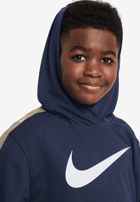 Tumši zils džemperis ar kapuci, izgatavots no mīkstas auduma, ar lielu baltu Nike logotipu un bēšas akcentu uz pleca. Ar kapuci.