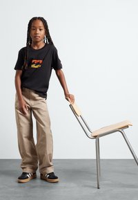 Camiseta negra con un gráfico multicolor, pantalones de pierna ancha beige y zapatillas negras con rayas blancas, de pie junto a una silla metálica minimalista.