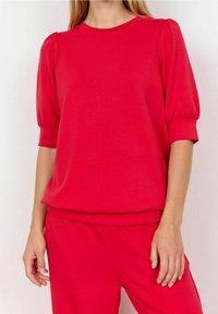 Suéter rojo con mangas cortas abullonadas y cuello redondo. Hecho de una tela suave con un corte holgado, presenta un puño y dobladillo acanalados.