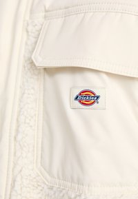 Giacca beige con uno strato interno testurizzato e soffice. Presenta una tasca a toppa e un logo Dickies colorato nella zona del petto.