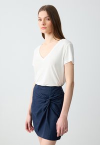 Weißes V-Ausschnitt Kurzarmoberteil, kombiniert mit einem dunkelblauen Rock mit knotigem Detail und glatter Textur. Einfaches und tailliertes Design.