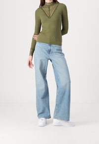 Olijfgroen lange mouw top met kantdetail, gecombineerd met lichtblauwe wijde jeans en witte sportschoenen. Vlakke stofstructuur en casual ontwerp.