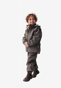 Vodoodporna rjav otroški jakna in hlače, ki vključujejo kapuco, elastične manšete in zadrge; oblečeno z rjavimi škornji.