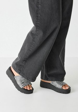 Chanclas de playa de patrón en blanco y negro con una banda ancha y suela gruesa y texturizada. Las bandas están hechas de un material suave y flexible.